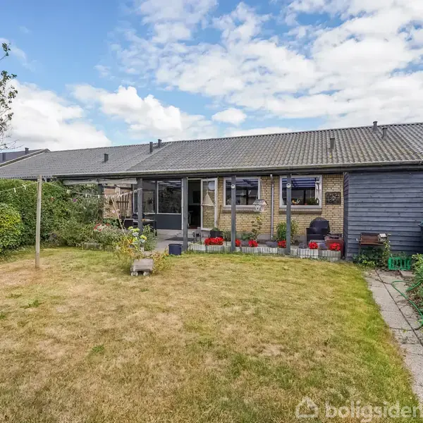 Et enfamiliehus med solarium, omkranset af en stor have med græsplæne og buske. Foran huset er en betonsti og nogle blomsterkrukker placeret på terrassen.