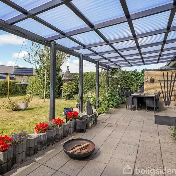 En overdækket terrasse med flisebelægning, hvor en ildskål står på gulvet. Omkring terrassen er der planter og blomster. Til højre er et bord og stole mod husets murstensvæg.