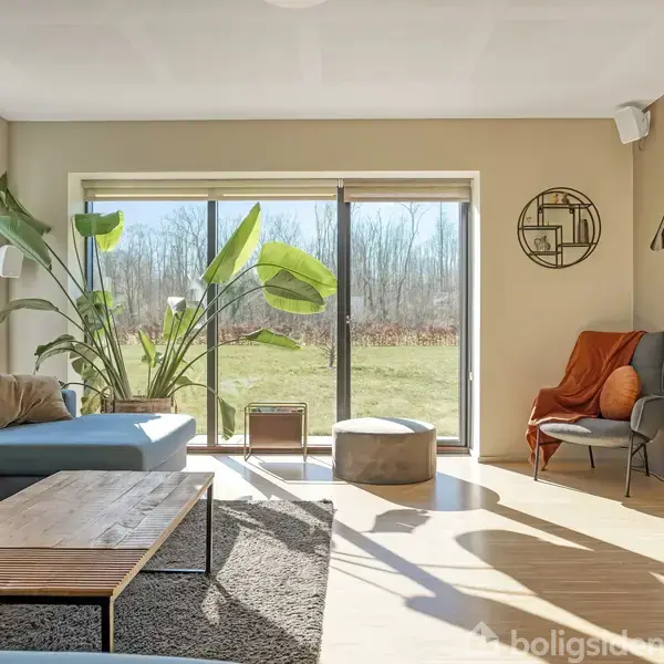 Stue med moderne, blå hjørnesofa ved glasdøre, der leder til haven. Sollys strømmer ind. Minimalistisk indretning med træbord, gulvtæppe, planter og lænestol med plaid i hjørnet.