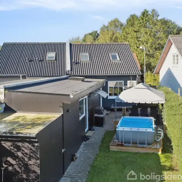 Et firkantet, fritstående hus har en græsplæne og en opstillet pool ved siden af. Omkringliggende have med hække og træer skaber en intim baggård.