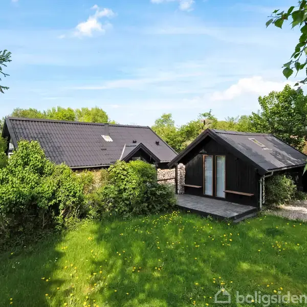 Et sort sommerhus med skråtag står stille i en frodig have omgivet af træer og buske under en blå himmel.