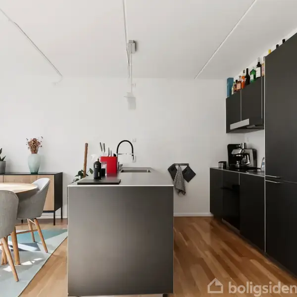Køkken med mørke skabe og en central ø, hvorpå der står en rød kedel. Ved siden af er et spisebord med grå stole. Rummet er lyst og moderne.