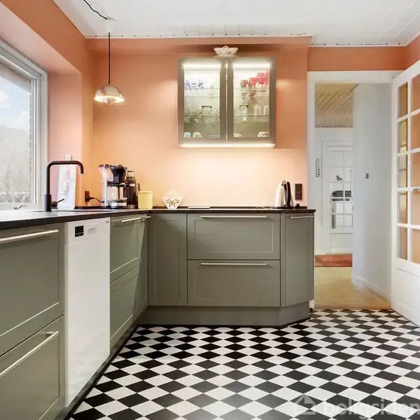 Køkken med grå skabe står i et hjørne, mens en lampe hænger over bordet. Fliser i sort-hvidt tern mønster dækker gulvet, omgivet af lyserøde vægge og store vinduer.