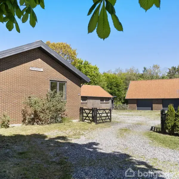 Brun murstensbygning står stille ved siden af grusvej, omkranset af grønne buske og træer. Baggrunden viser en anden bygning med rødt tag. Ingen synlig tekst.