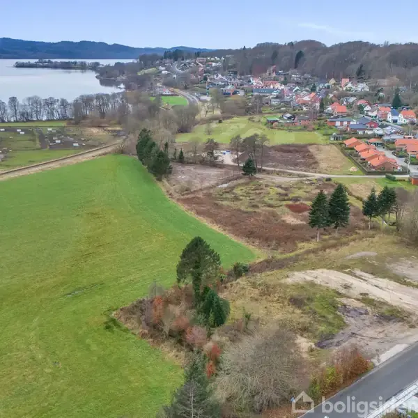 Landskab med marker og en sø i baggrunden. En landsby ligger til højre med rødt tagede huse. En vej løber langs højre side, flankeret af træer og buske.