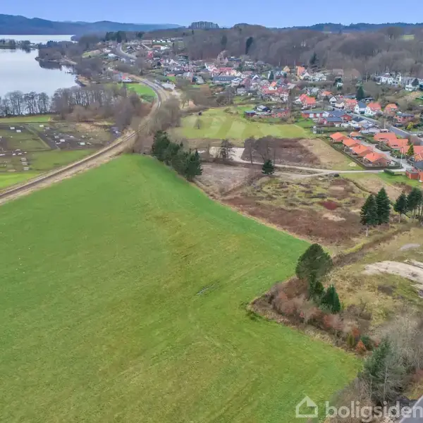 Grønt landskab med spredte træer strækker sig mod en sø til venstre. Til højre ses en lille by med røde hustage, omgivet af veje og en skovklædt baggrund.