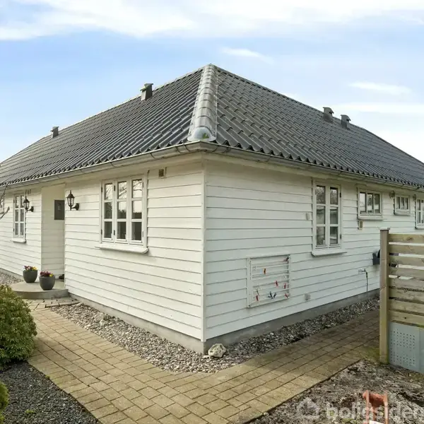 Hvidmalet hus med sort tag og flere klassiske vinduer. Det står på en grund med flisebelagt gangsti og grus, omringet af træhegn og buske, under en klar himmel.