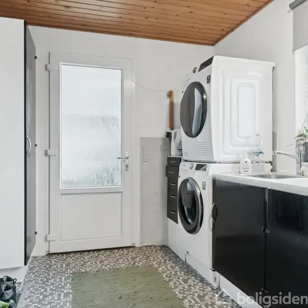 En vaskemaskine og tørretumbler er stablet ved siden af en vask med planter på bordet; stuebordet er ved en glasdør. Gulvet har mønstret belægning.