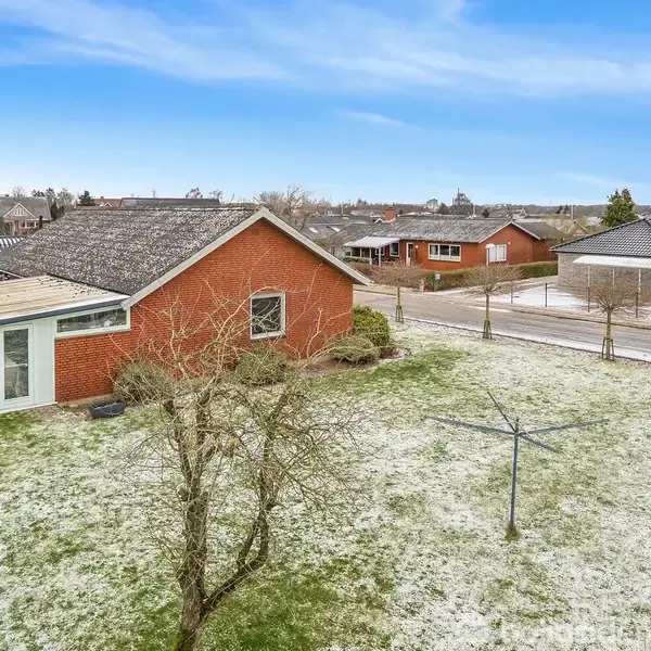 Rødt hus står stille på en let sneklædt græsplæne med et tørrestativ i forgrunden. Flere huse ses i baggrunden under en klar, blå himmel.