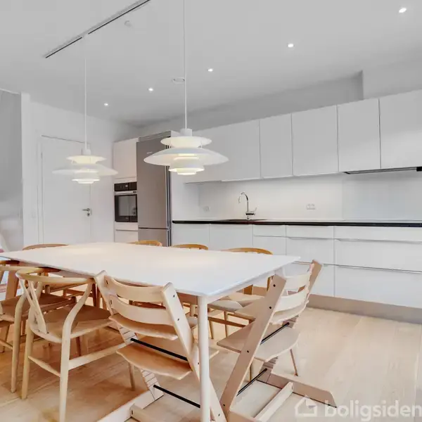 Et moderne køkken har et stort spisebord med træstole. Over bordet hænger tre hvide lamper. Køkkenet har hvide skabe og en indbygget ovn. En trappe ses til venstre.
