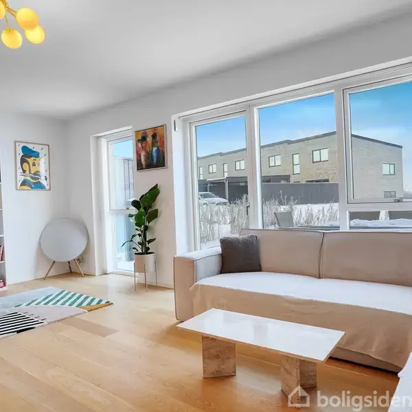 Beige sofa ved et stort vindue i en moderne stue, med et marmorbord foran. Bogreol fyldt med bøger på venstre væg; lys træparketgulv. Rund lampe hænger ned fra loftet.