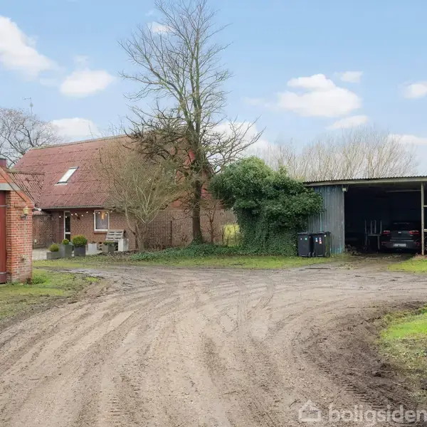 En grusvej fører til en gårdsplads med en åben garage, hvor en bil står parkeret. Til venstre ses en rød murstensbygning og et hus med skråt tag. Træer omgiver området.