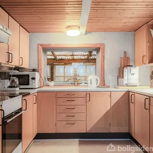 Køkken med træskabe og hvid bordplade, udstyret med en komfur, ovn, mikroovn og elkedel. Et vindue over vasken åbner ud mod en overdækket terrasse med trækonstruktion.