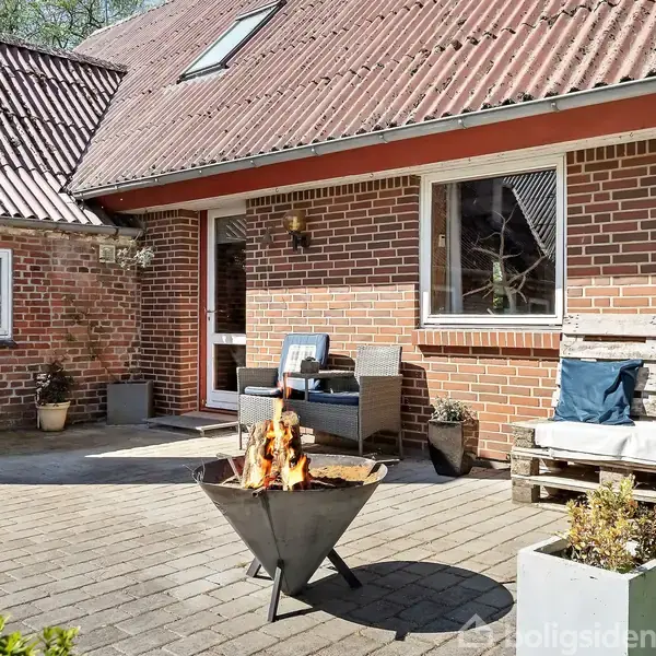 Bålsted brænder i en trekantet beholder på en brolagt terrasse foran et murstenshus. To stole og planter er placeret omkring bålstedet i et hyggeligt gårdmiljø.
