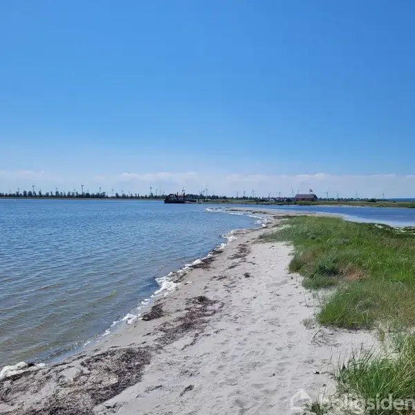 En kystlinje strækker sig langs en sandstrand med blåt vand til venstre og grønt græs til højre under en klar blå himmel. Holzbygninger ses i det fjerne.