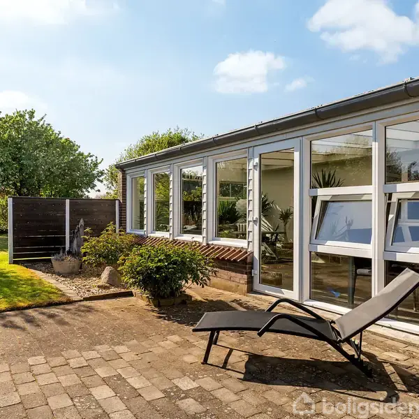 En solrig terrasse med en liggestol foran et moderne hus med store vinduer. Haven omkring har grønne træer og buske, samt en flisebelagt gangsti.