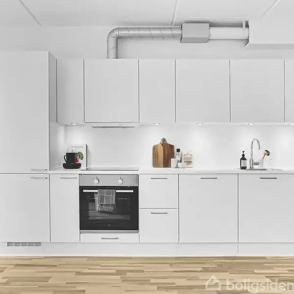 Hvidt køkken med integreret ovn og komfur, stående på trægulv. Vægskabe og hylder med dekorationer, skærebrætter, en plante og kurve tilføjer detaljer. Ventilation over skabene.