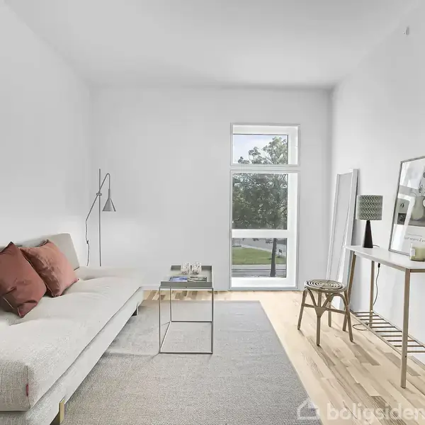 Sofa med røde puder står i en lys stue. En moderne gulvlampe ved siden af, og et bord med dekorationer står ved vinduet med udsigt til træer.