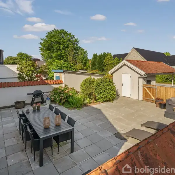 En terrasse med et sort spisebord og stole er omgivet af planter og hegn. Grill står på flisebelagt område, under en blå himmel med hvide skyer.