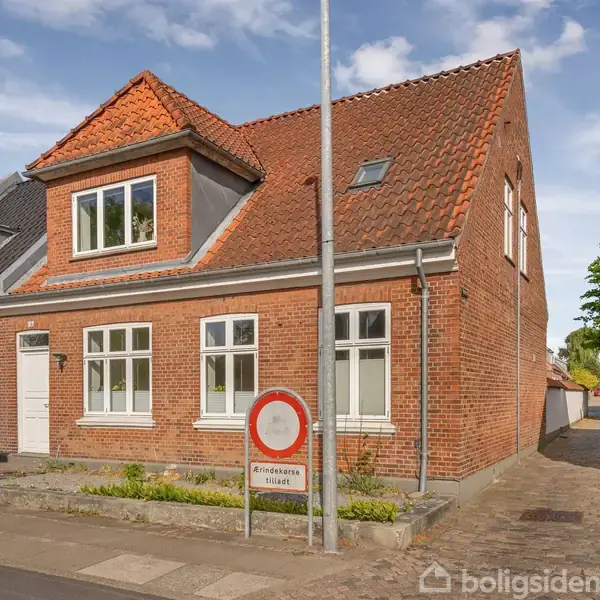 En murstensbygning med rødt tegltag og flere vinduer står langs en stille gade. Foran huset er der et skilt med teksten: "Gennemkørsel forbudt." Omkring er der andre huse og træer.