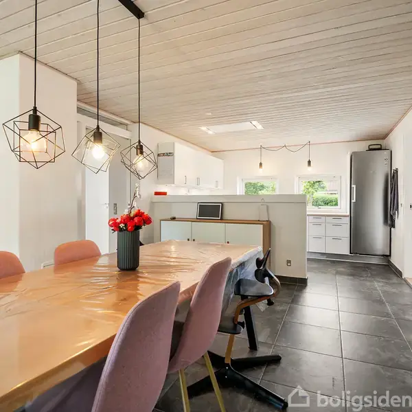 En spisestue har et langt bord med lyserøde stole og en vase med røde blomster. Tre moderne lamper hænger over bordet. Køkkenet i baggrunden er lyst og moderne.