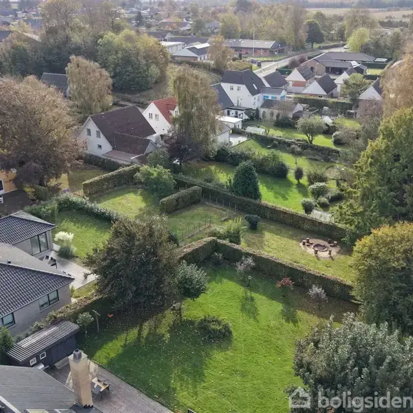 En række huse med farverige tage omkranser en række haver og grønne områder omgivet af træer og buske i et beboelseskvarter.