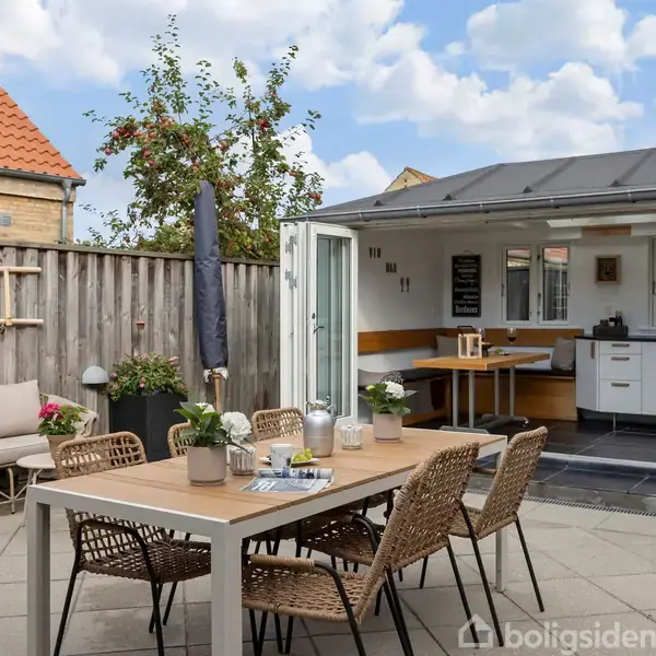 En udendørs spisestue med bord og seks stole står på en terrasse ved siden af et åbent skur. Skurets døre er åbne, og der er et lille køkken indeni.