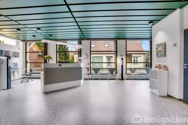 Reception med skranke står ubenyttet i en moderne klinik. Stole står ved vindue i en lys, indbydende ventesal med udsigt til traditionelle huse udenfor.