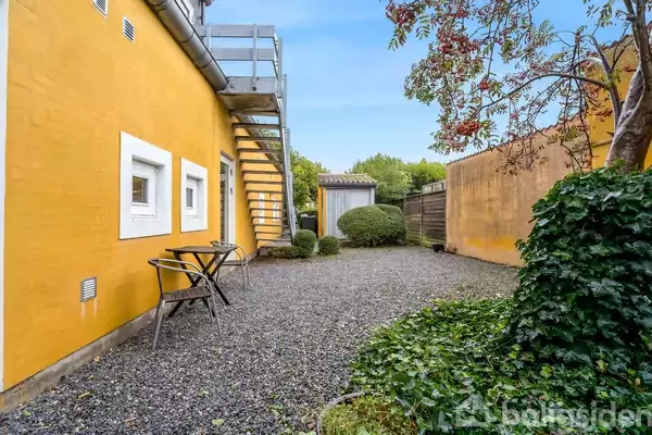 Et bord med to stole står foran en gul bygning med vinduer og en udendørs trappe. Grusbelagt gårdhave omkranset af buske og træer.