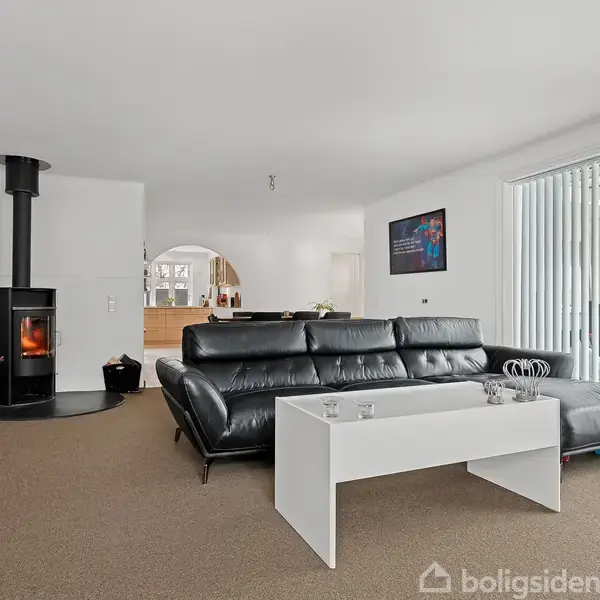 Pejs opvarmer en stue med en sort L-formet sofa og et hvidt sofabord. Kort på væggen, store vinduer med lodrette persienner. Hyggelig, moderne indretning.
