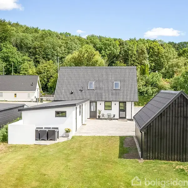 Hus med sort tag og vinduer står på et velholdt grønt græsareal, omgivet af skov og andre bygninger. Terrasse møbler foran huset indikerer en afslappende udendørs opholdsplads.