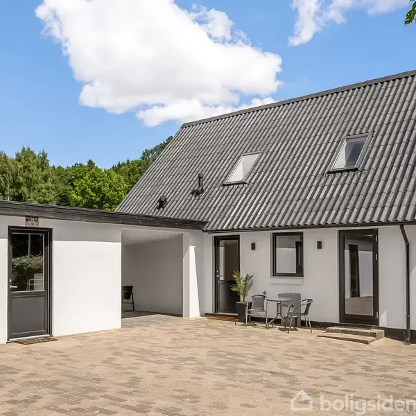 En hvidmalet hus med sort tag står stille på en flisebelagt gårdsplads. Der er en lille terrasse med to stole. Omgivelserne er skovklædte med blå himmel og få skyer.