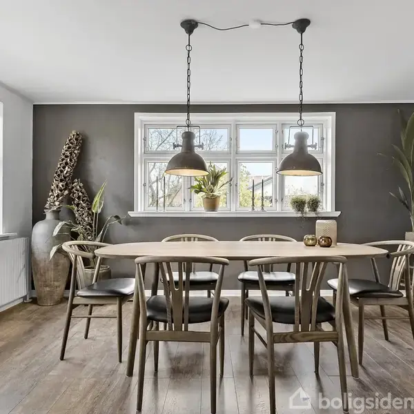 En stor spisebord i træ omgivet af seks stole står i en lys spisestue. Flere planter og to loftlamper dekorerer rummet, mens vinduet giver udsyn til naturen udenfor.