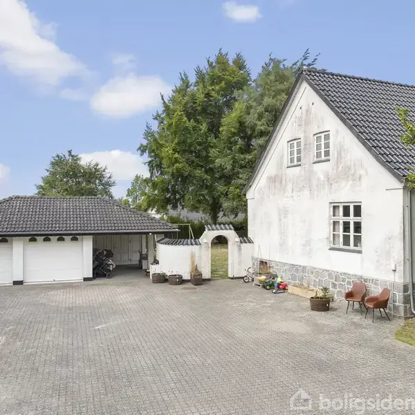 Et hvidt hus med sort tag står ved siden af en garage med to porte. Foran er en stor brolagt gårdsplads, omgivet af grønne træer og buske.
