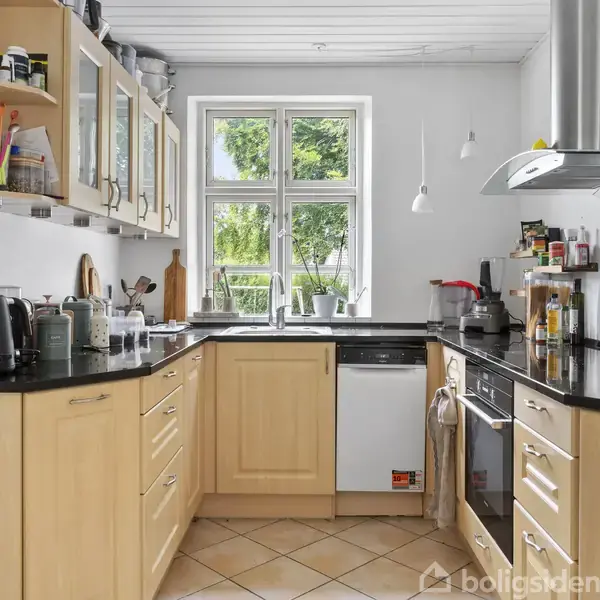 Køkken med lyse træskabe og sort bordplade. Et vindue i midten slipper lys ind. Der står husholdningsapparater og frugtskåle. Fliser på gulvet og hvidt loft fuldender rummet.