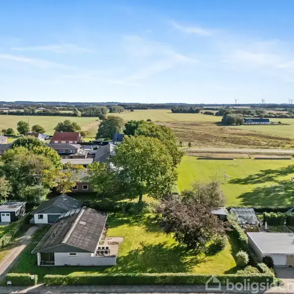 Et rækkehus er omgivet af træer og haver i et landligt område. I baggrunden strækker markerne sig ud mod horisonten under en blå himmel.
