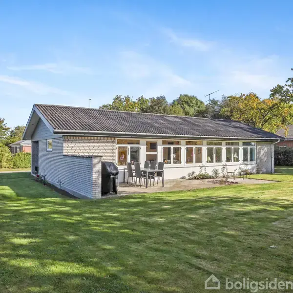 Et enfamiliehus af mursten står i en velplejet have med en terrasse foran. Terrassebord og stole er opstillet, omgivet af hæk og træer under en klar blå himmel.