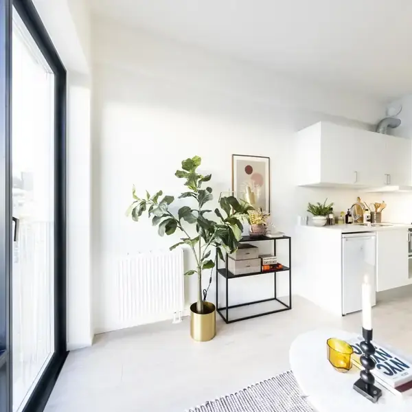 En stue med et hvidt køkkenhjørne, grøn plante i gulvkrukke, og sofabord med lys og gul dekoration. Store vinduer til venstre giver naturligt lys.