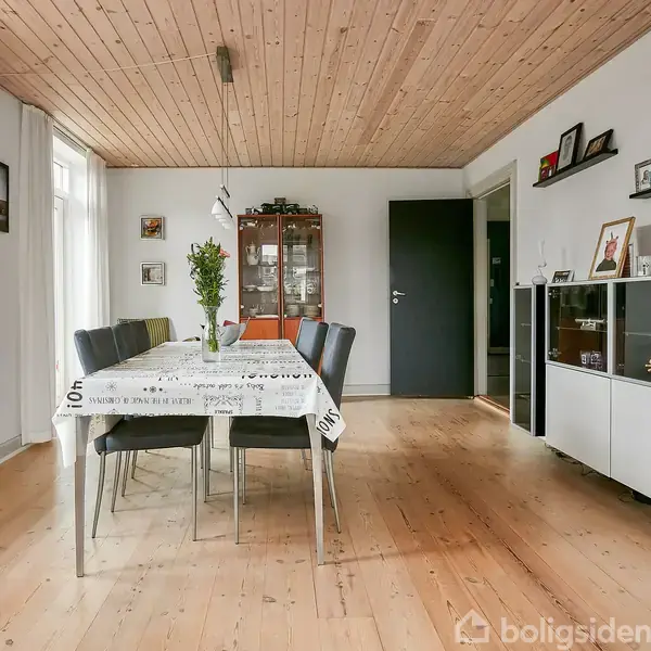 Spisebord dækket med dug står i et rum med trægulv og loft. Et skab med glaslåger står i hjørnet. Vægge har forskellige billeder og en reol med genstande.