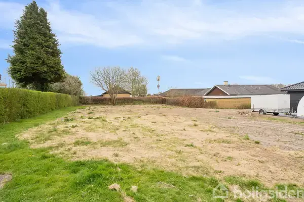 En tom byggegrund dækket med tør jord og spredt græs, omgivet af hække og træer. Grunden ligger i et beboelseskvarter med huse i baggrunden under en blå himmel.