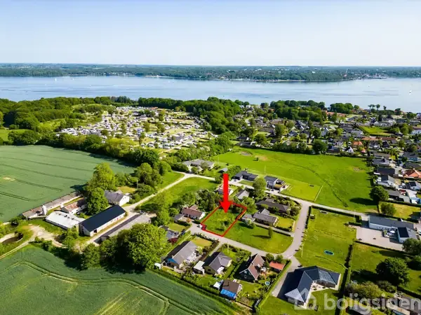 En rød pil peger på en grøn grund omgivet af huse i et villakvarter med græsplæner, træer og en fjord i baggrunden.