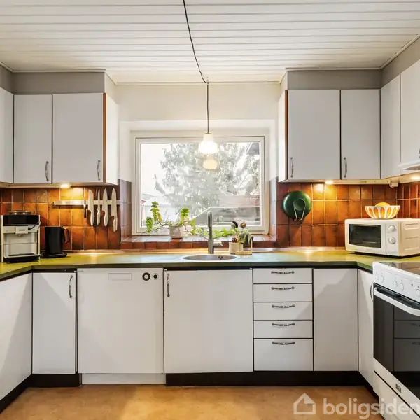 Hvidt køkken med grønne bordplader. Kaffemaskine, knive og gryder hænger på væggen. Midt i rummet er et vindue med planter. Orange-brune fliser danner baggrunden.