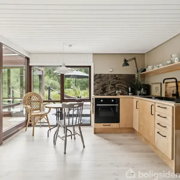 Køkken med en træbordplade, indbygget ovn, og åbne hylder, forbundet til spisestue med glasvægge. Udsigt til terrasse med havemøbler og grønne omgivelser.