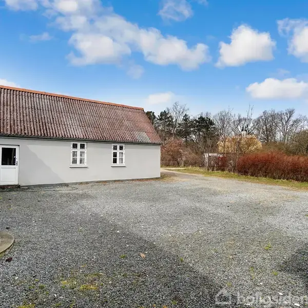 Et gråmuret hus med rødt tegltag står stille på en grusbelagt gårdsplads. I baggrunden ses buske og træer under en delvist skyet blå himmel.