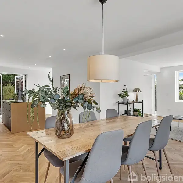 Et lyst rum med spisebord i træ og grå stole. En loftlampe hænger over bordet, dekoreret med en vase med blomster. Stuen i baggrunden har sofa, lænestol og vinduer med lysindfald.