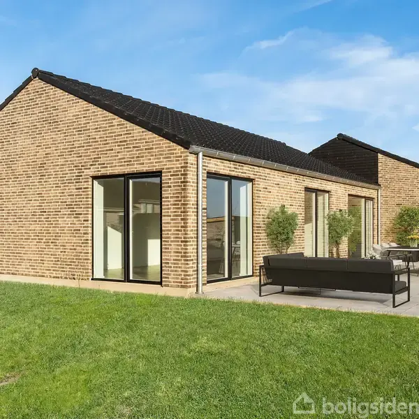 En moderne villa med murstensfacade og store vinduer, placeret i en have med græs. En terrasse med sofa og spisebord er opstillet foran huset.