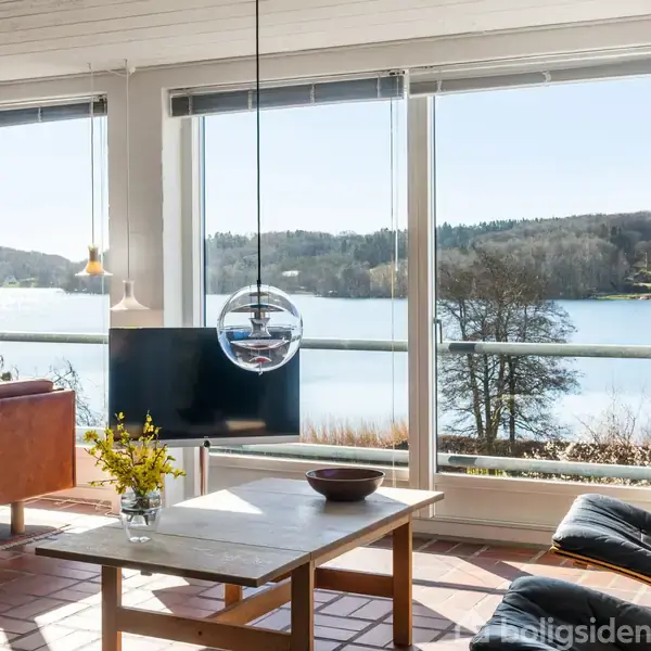 Stue med stort vinduesparti viser udsigt til sø og skov. Sollys strømmer ind, rammer sofaen, en lænestol, et sofabord med en vase. Billede på væggen.