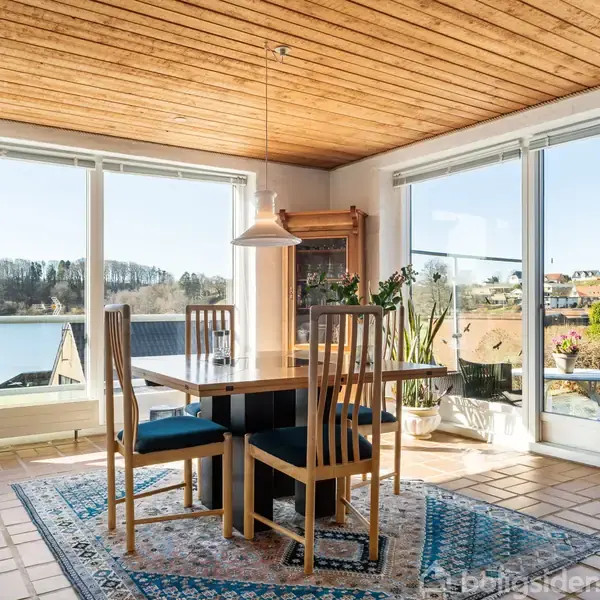 Spisebord med fire stole står i en lys spiseplads. Store vinduer på begge sider afslører udsigt til en sø og bakker med huse. Tæppe ligger under bordet.