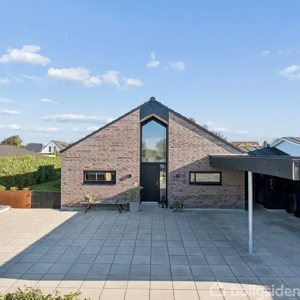 En moderne villa med trekantet facade og stort vindue står på en flisebelagt gårdsplads. Til højre er en overdækket carport, og grønne hække omkranser ejendommen.