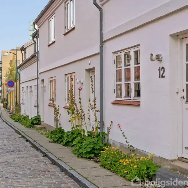 Hvide byhuse med nummer 12 ses langs en brostensbelagt gade. Der vokser blomster og planter langs husmurene. En bil er parkeret til venstre.